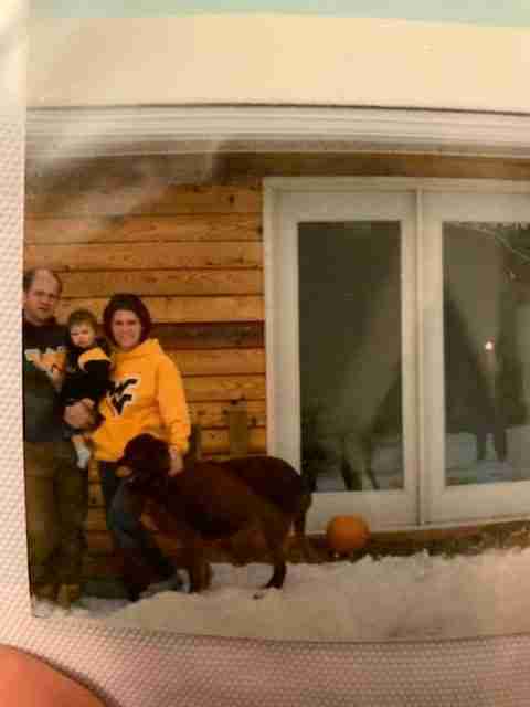 Man, and woman holding a baby in WV hoodies with an irish setter standing on a porch with snow by glass doors on a wooden house.