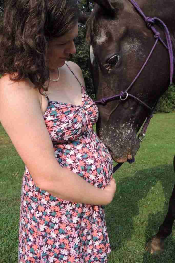 White woman with shoulder length brown hair; white pearl necklace; pink, white, and maroon flower sundress with her arm wrapped around her pregnant belly. Brown horse with a white star on her head; and white spots on her muzzle; wearing a purple rope halter. Horse has her eye half closed and is nuzzling womans pregnant belly. They are standing in bright green short grass.