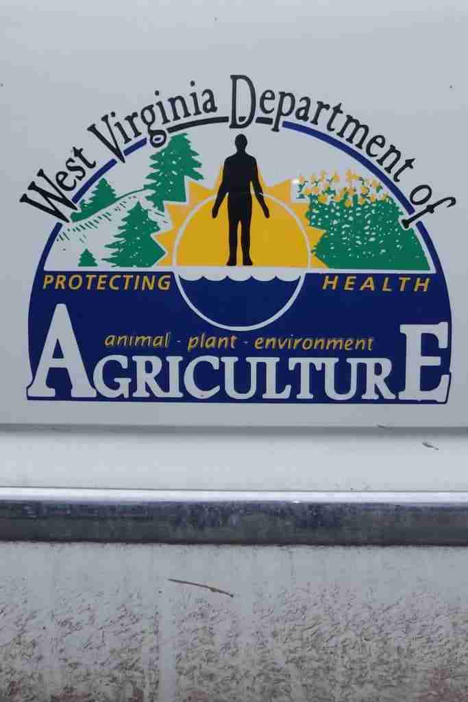 West Virginia Department of Agriculture Logo. West Virginia Department of written over green pine tree graphics and a human silhouette over a sun with green and yellow graphic flowers to its left. Under this is written Protecting Health in yellow on a dark blue background. Under is written: animal-plant-environment. And at the very bottom Agriculture is written in white.