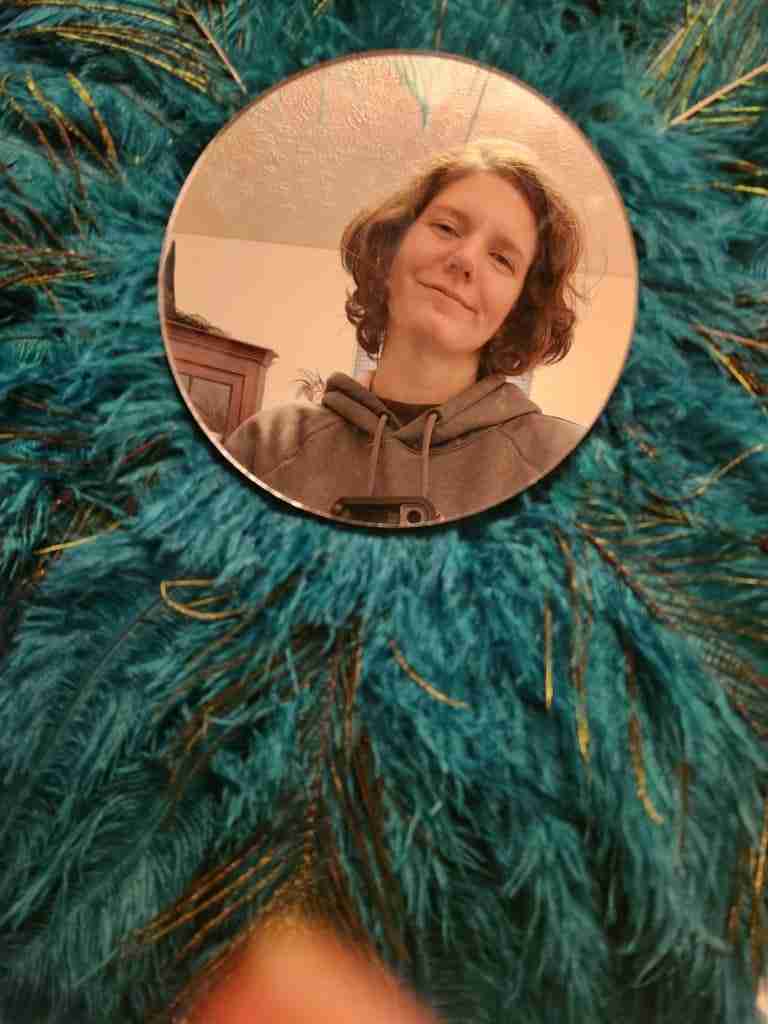 White woman with brown hair parted at the side and cut just under jaw length; wearing a grey hoodie. Smiling with mouth closed pictured from the shoulders up, looking out from a round mirror that has blue green feathers radiating out from behind it.