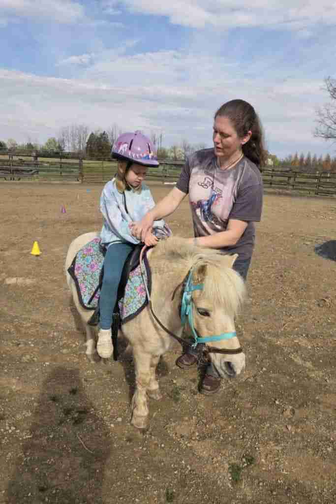 White woman with dark brown hair in ponytail wearing grey short sleeve t-shirt with cream front and blue horse on it blue jeans; and dark brown boots helping during a child's horse riding lesson on a cream pony wearing a blue bitless bridle and brown leather halter. Child is wearing a purple helmet; blue jeans; light blue and white long sleeve shirt and white boots; sitting on black english saddle with blue and purple saddle pad. Standing in dirt arena with a yellow cone; purple cone; and red cone on the ground behind them for fun horse games for kids surrounded by 3 board wood fences
