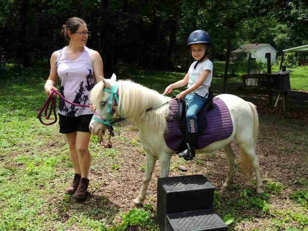 White woman dark brown hair in a bun (Zuri). Wearing black shorts; brown leather Ariat boots; sleeveless shirt light purple with abstract black mottling. Leading a white pony wearing a blue bitless bridle and reins; purple LeMieux saddle pad; black Wintec saddle. 4-year-old child riding in short green grass with a 2 step black mounting block on the ground.