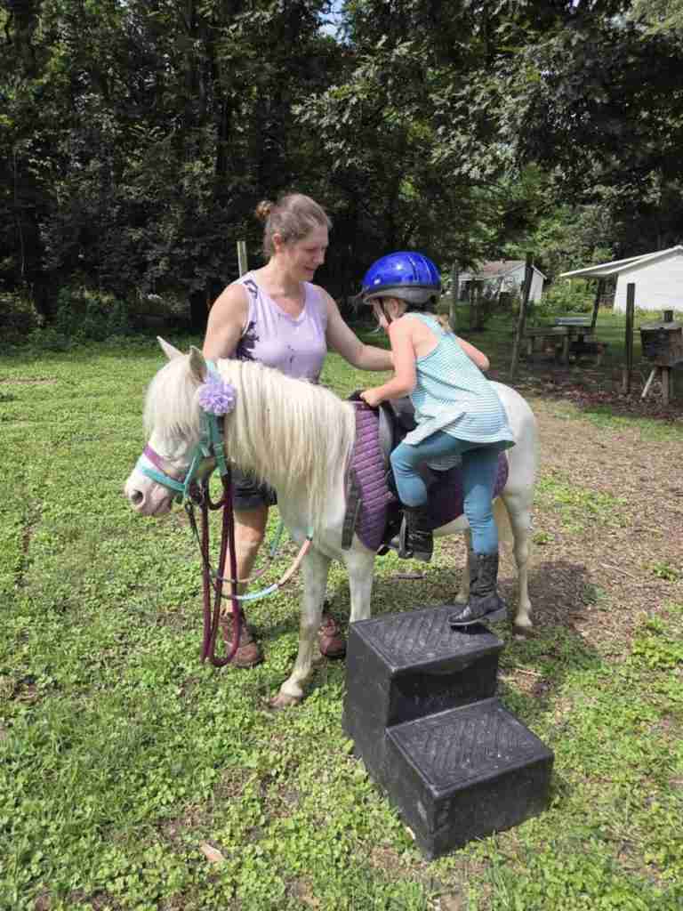 White woman dark brown hair in a bun (Zuri). Wearing black shorts; brown leather Ariat boots; sleeveless shirt light purple with abstract black mottling. Standing by a white pony wearing a blue bitless bridle with a purple flower by her ear; purple LeMieux saddle pad; black Wintec saddle. 4-year-old child with her foot in the stirrup on a black 2 step mounting block in short green grass.