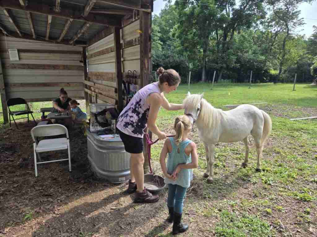 White woman dark brown hair in a bun (Zuri). Wearing black shorts; brown leather Ariat boots; sleeveless shirt light purple with abstract black mottling. Standing in front of white pony wearing a purple halter. 4-year-old child riding standing facing the pony (Zony). PAL Around Pony School setup behind them with a mother and child working at the kids picnic table.