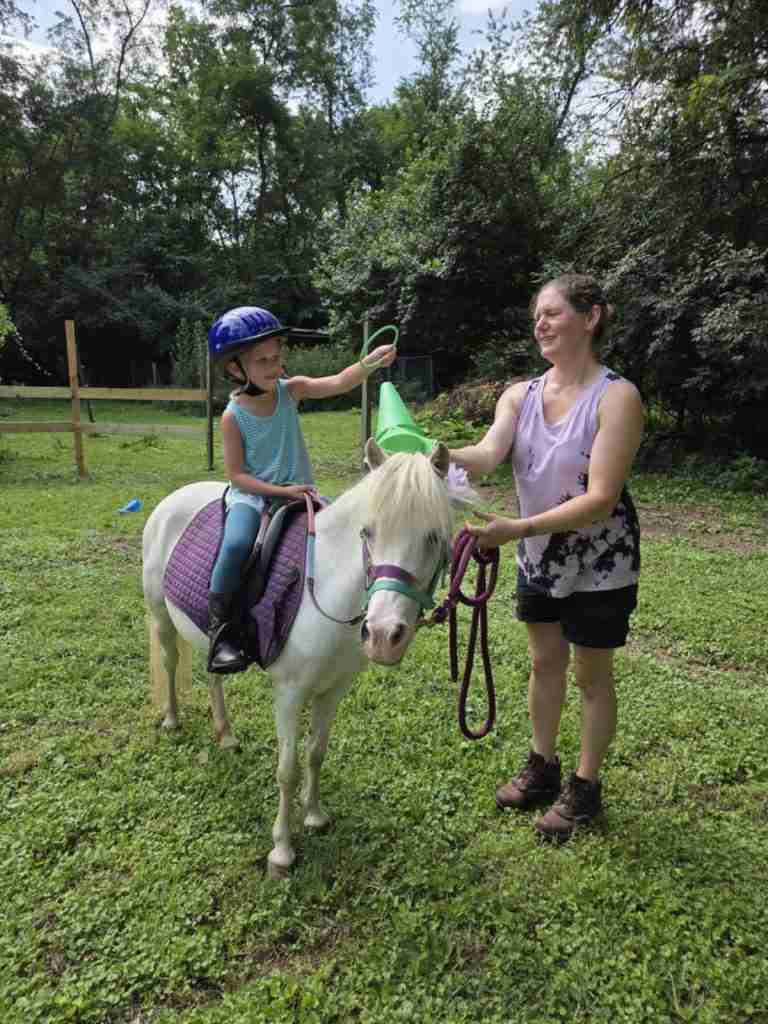 White woman dark brown hair in a bun (Zuri). Wearing black shorts; brown leather Ariat boots; sleeveless shirt light purple with abstract black mottling. Standing next to a white pony wearing a blue bitless bridle and reins; purple LeMieux saddle pad; black Wintec saddle. 4-year-old child riding in short green grass reaching to put a ring over a cone during a pony riding lesson.
