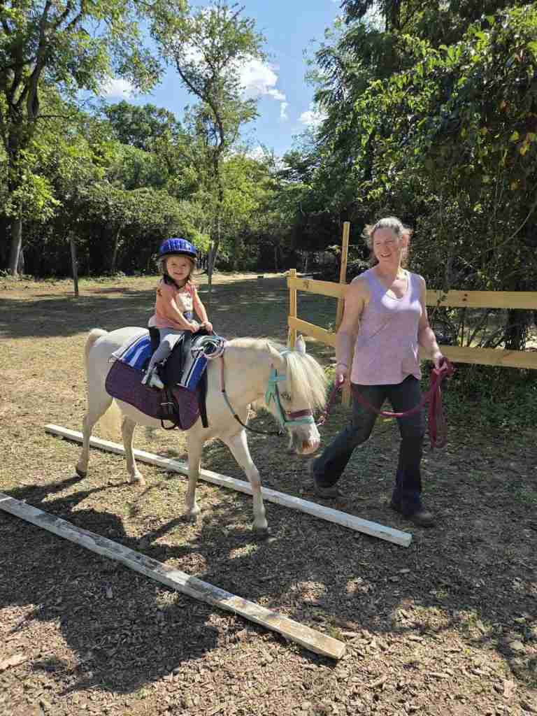 White woman dark brown hair in a bun (Zuri). Wearing black shorts; brown leather Ariat boots; sleeveless shirt light purple with abstract black mottling. Leading a white pony wearing a blue bitless bridle and reins; purple LeMieux saddle pad; black Wintec saddle. 2-year-old child riding in a dirt arena walking between two white poles.