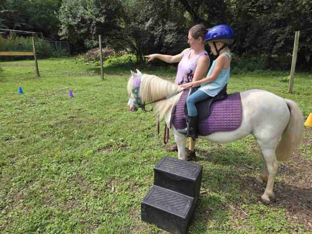 White woman dark brown hair in a bun (Zuri). Wearing black shorts; brown leather Ariat boots; sleeveless shirt light purple with abstract black mottling. Pointing to show student where to ride. Standing next to a white pony wearing a blue bitless bridle and reins; purple LeMieux saddle pad; black Wintec saddle. 4-year-old child riding in leggings, and boots which are good beginner horseback riding clothes for kids. On short green grass with a 2 step black mounting block on the ground.