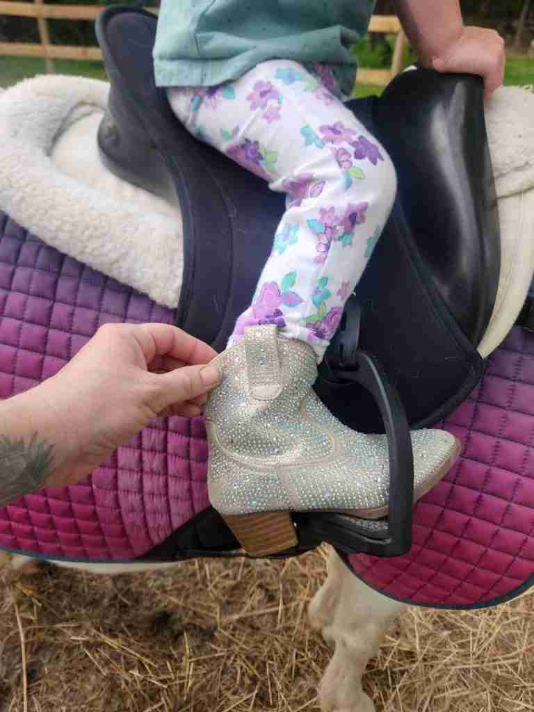 Side view of a cowboy boot with too high of heel in a stirrup with a purple saddle pad and black saddle behind it.