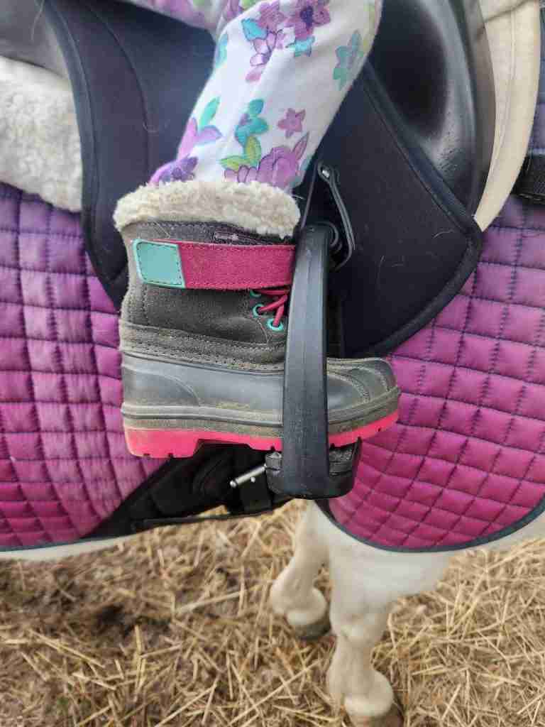 Cat & Jack snow boot in a stirrup with a purple saddle pad and black saddle behind it.
