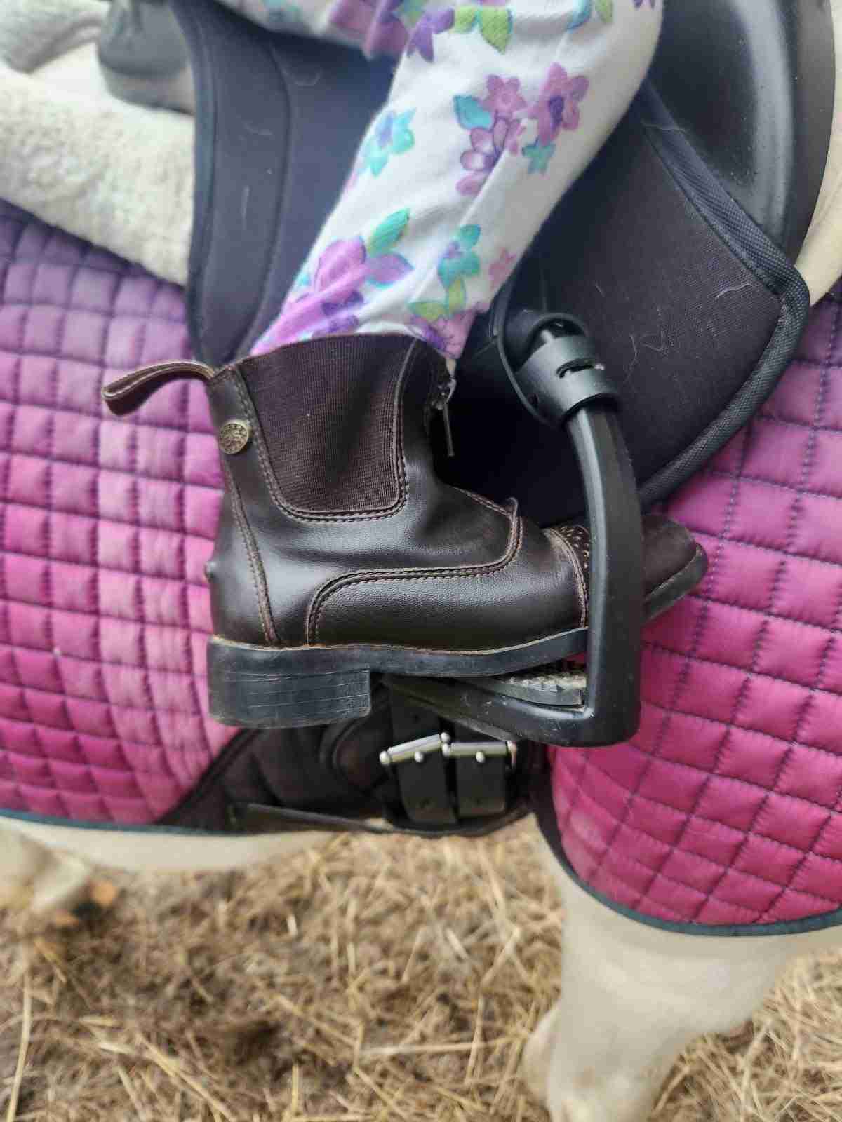 Children's horseback riding paddock boot in a stirrup with a purple saddle pad and black saddle behind it.