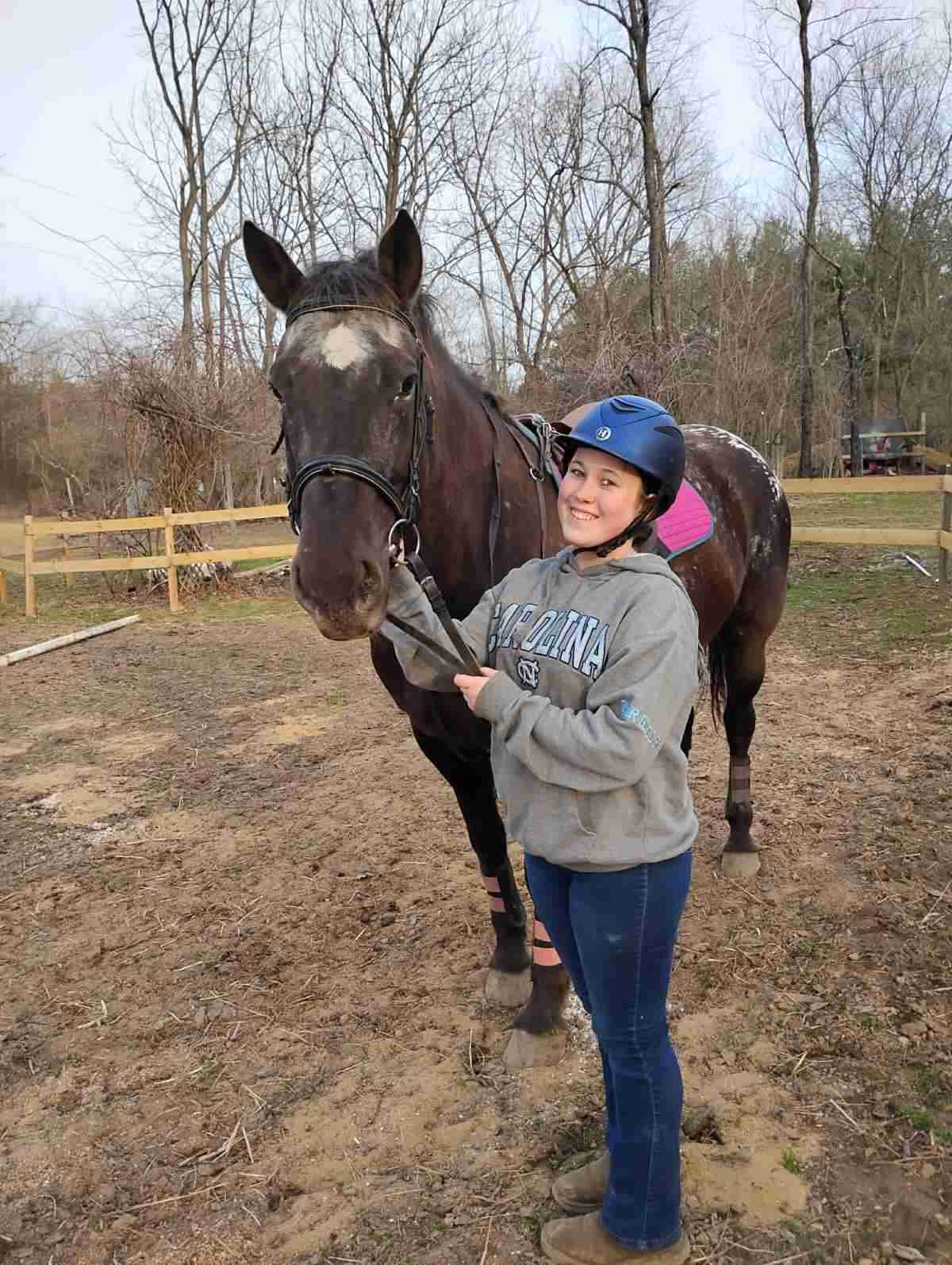 horse riding lessons for kids. 14 year old kid with blue riding helmetholding the reins to a brown horse with white star standing in a grass riding arena.